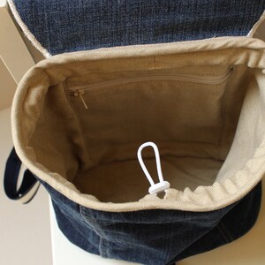 May include: A denim backpack with a beige drawstring top and a zippered pocket inside. The backpack has a white drawstring cord.