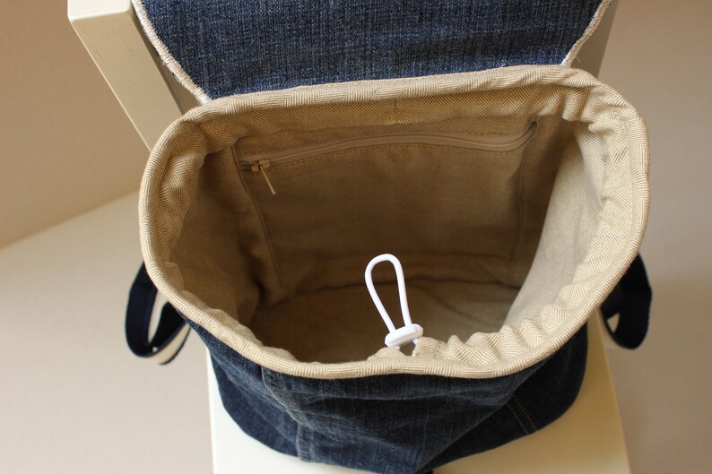 May include: A denim backpack with a beige drawstring top and a zippered pocket inside. The backpack has a white drawstring cord.