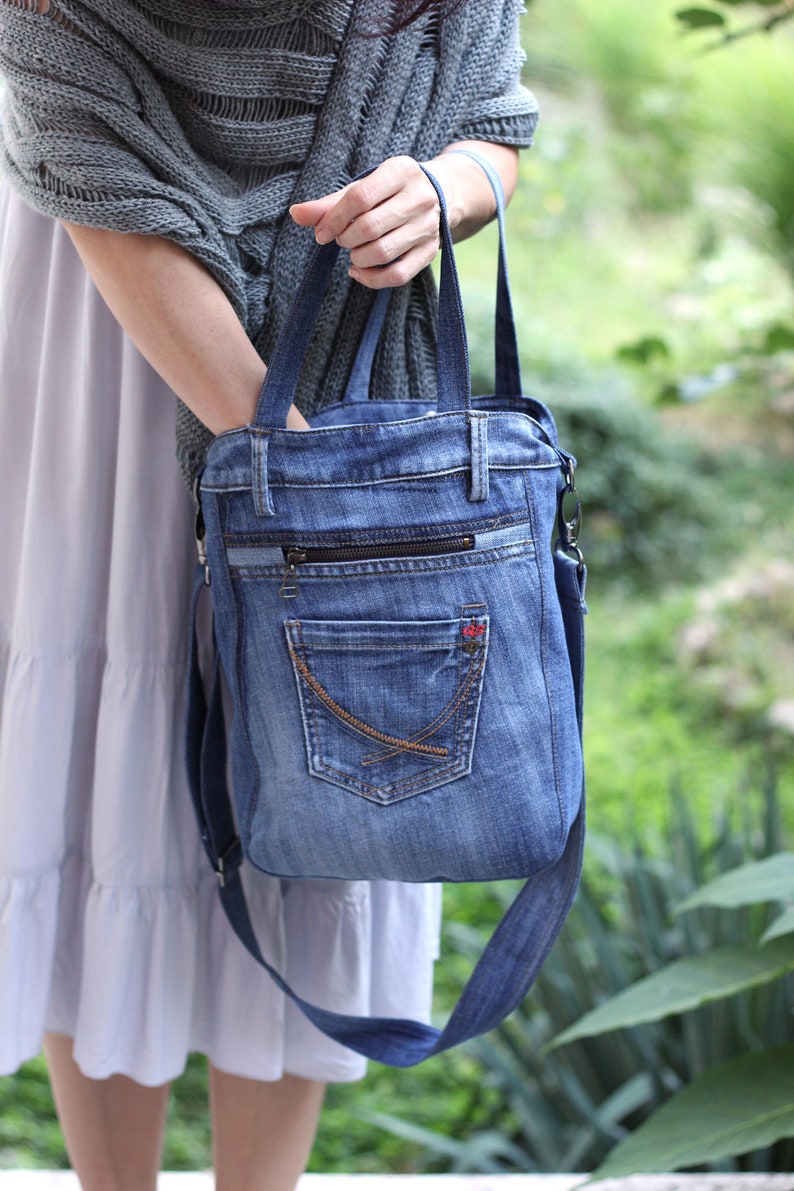 May include: A denim tote bag with a zippered pocket and a large front pocket. The bag has a long strap that can be worn over the shoulder.