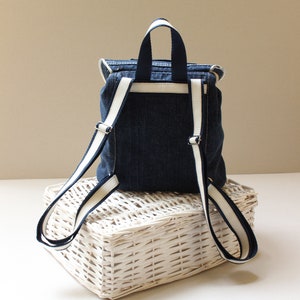 May include: A small denim backpack with white trim on the straps. The backpack is sitting on a white wicker basket.