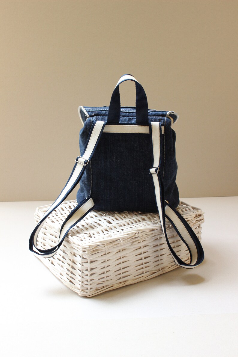 May include: A small denim backpack with white trim on the straps. The backpack is sitting on a white wicker basket.