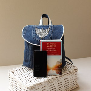 May include: A blue denim backpack with a white trim and a rhinestone embellishment. The backpack is sitting on a white wicker basket with a black smartphone and a red and white book titled "Le Pouvoir de l'Intention" by Dr Wayne W. Dyer.