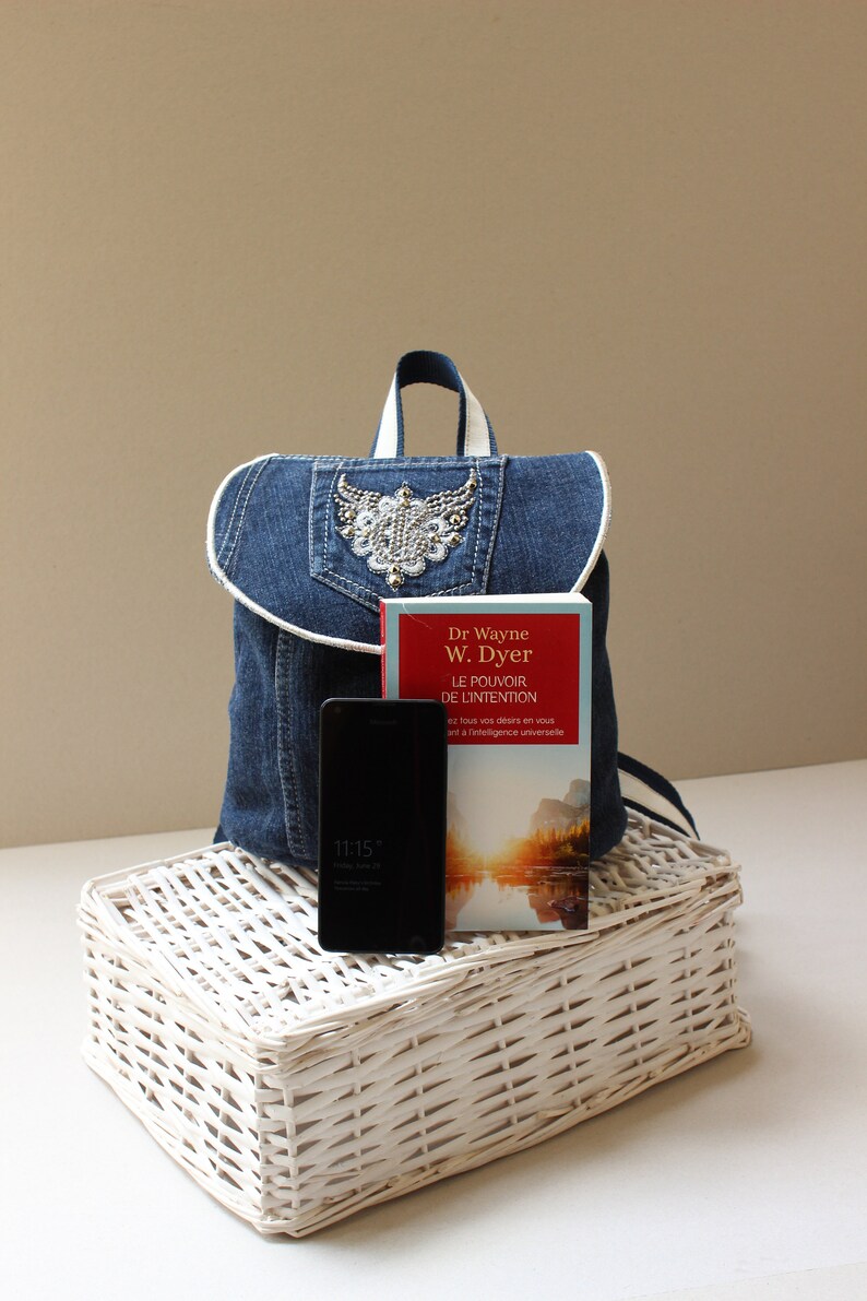 May include: A blue denim backpack with a white trim and a rhinestone embellishment. The backpack is sitting on a white wicker basket with a black smartphone and a red and white book titled "Le Pouvoir de l'Intention" by Dr Wayne W. Dyer.