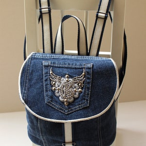 May include: A denim backpack with white straps and a decorative pocket featuring a beaded design. The backpack is sitting on a white chair.