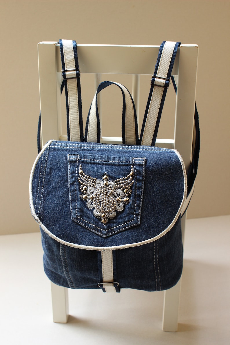 May include: A denim backpack with white straps and a decorative pocket featuring a beaded design. The backpack is sitting on a white chair.