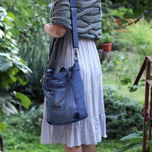 May include: A woman wearing a gray knitted sweater and a white skirt is carrying a blue denim tote bag with a long strap. She is wearing blue wedge sandals.