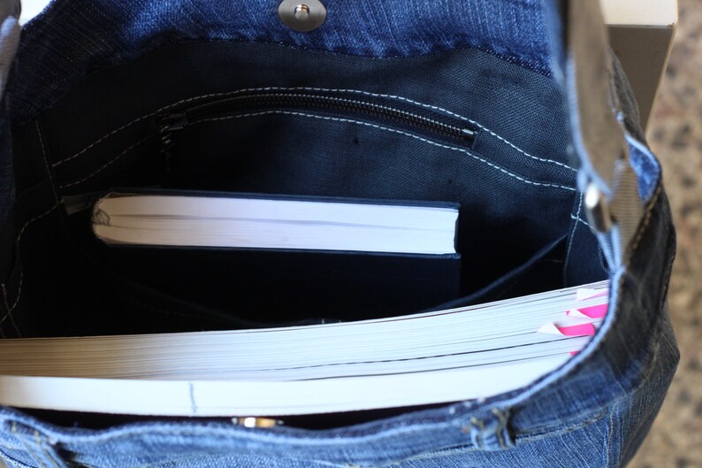 May include: A blue denim tote bag with a zippered pocket and a white book inside. The bag is open and the book is visible.