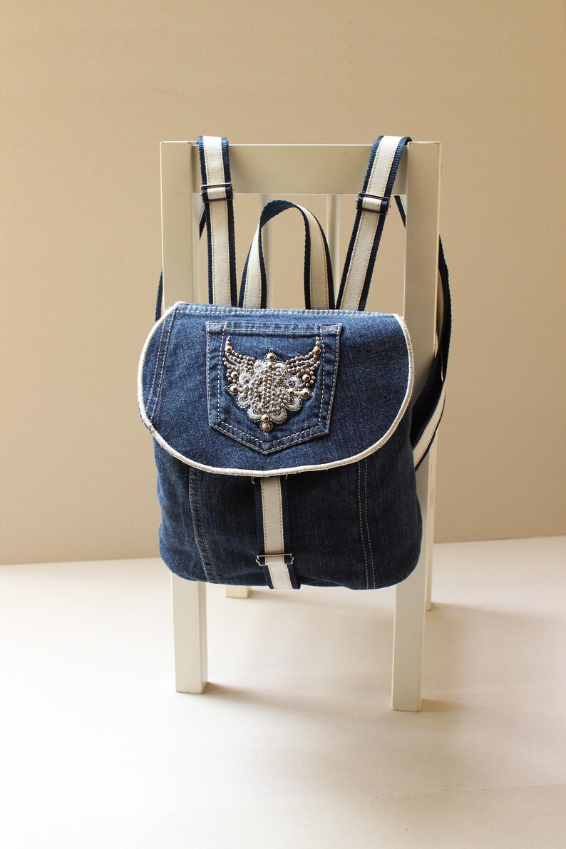 May include: A blue denim backpack with white straps and a decorative embellishment on the front pocket.
