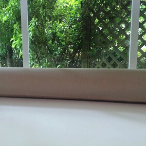 May include: A close-up of a beige window valance with a view of green foliage and a wooden lattice fence through the window.