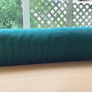 May include: A rolled-up bolt of dark teal fabric, likely velvet or a similar material, rests on a light-colored wooden surface. The fabric is a solid color, and the roll is positioned horizontally. The background includes a lattice fence and greenery.