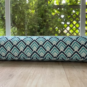 May include: A long, cylindrical pillow with a repeating geometric pattern in shades of teal and navy blue on a white background. The pillow is resting on a light-colored wooden floor, with a window and greenery visible in the background.