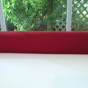 May include: A long, cylindrical, solid red bolster pillow. The pillow is made of a soft, plush material and is resting on a white surface. The background features a window with greenery.