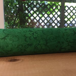 May include: A rolled-up length of green fabric with a dark green, swirling floral pattern. The fabric is resting on a wooden surface, with a window and outdoor greenery visible in the background.