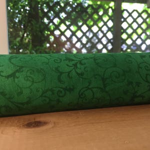 May include: Green fabric with a swirling, vine-like pattern. The fabric is rolled up and sitting on a wooden surface.