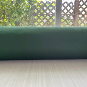 May include: A rolled-up, dark green fabric, possibly velvet or a similar material, rests on a light-colored wooden surface. The background features a window with a lattice design and lush green foliage.