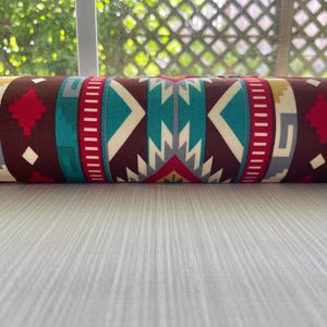 May include: Rolled fabric with a Southwestern pattern. The design features geometric shapes in teal, red, brown, and cream. The fabric is rolled up, showcasing the repeating pattern. The overall aesthetic is reminiscent of Native American textiles.