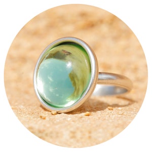 Op de afbeelding: Een zilveren ring met een grote, ronde, groene glazen steen. De steen is reflecterend en toont een bewolkte lucht.