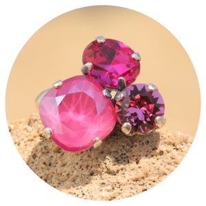 Può includere: Un anello d'argento con tre vivaci pietre rosa. La pietra più grande è a taglio cuscino, quella centrale ovale e la più piccola rotonda. L'anello è appoggiato su una superficie beige e strutturata, che mette in risalto il design e il colore del gioiello.