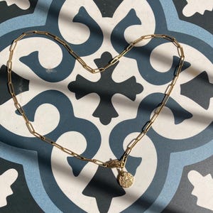 Könnte beinhalten: Eine goldene Kettenkette mit einem kleinen, runden, strukturierten Anhänger. Die Kette liegt in Form eines Herzens auf einer blauen und weißen gekachelten Oberfläche.