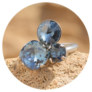 Puede incluir: Un anillo plateado con tres cristales grandes, facetados y de color azul. Los cristales están engastados en un diseño de racimo, con una piedra redonda y dos piedras cuadradas. El anillo se muestra sobre una superficie texturizada de color beige.