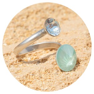 Op de afbeelding: Een zilverkleurige ring met twee grote, ronde, gefacetteerde edelstenen. Eén is helder, de andere lichtblauw. De band van de ring buigt zich om de tweede edelsteen te ontmoeten, waardoor een uniek ontwerp ontstaat.