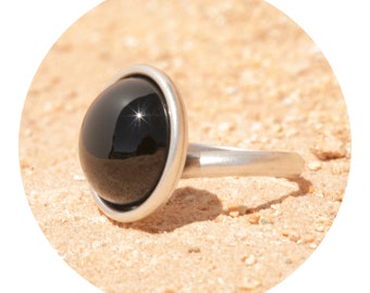 Op de afbeelding: Een zilveren ring met een zwarte, ronde edelsteen. De edelsteen is gezet in een zilveren bezel en reflecteert licht.