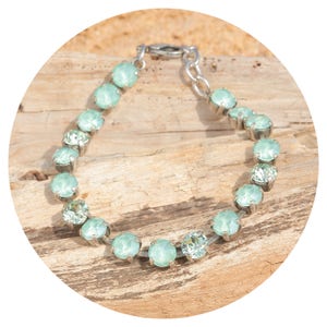 Op de afbeelding: Een zilveren armband met ovale, lichtgroene edelstenen. De armband is gezet op een zilveren ketting en sluiting. De edelstenen hebben een zachte, pastelgroene kleur. De armband wordt getoond op een houten oppervlak.