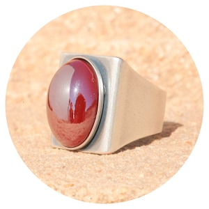 Könnte beinhalten: Ein silberner Ring mit einem großen, runden, dunkelroten Edelstein, der in einer quadratischen Fassung sitzt.