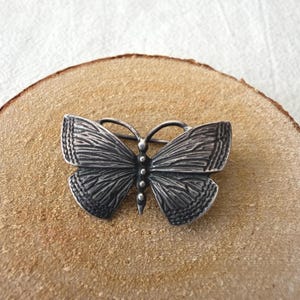 Op de afbeelding: Een zilverkleurige vlinderbroche met een gedetailleerd, getextureerd vleugelpatroon met donkere verdiepte lijnen en een centraal lichaam bestaande uit een verticale rij kleine, afgeronde bolletjes. De vleugels hebben een geschulpte randafwerking.
