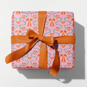 May include: A wrapped gift box with pink wrapping paper patterned with orange bows and green vines. It's tied with a wide orange satin ribbon, knotted in a bow on top. The gift is presented on a white surface.