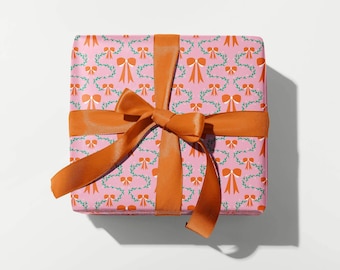 Op de afbeelding: Een ingepakte geschenkdoos met roze cadeaupapier met een patroon van oranje strikken en groene slingers. De doos is vastgebonden met een breed, oranje satijnen lint, dat een strik vormt. Het cadeau ligt op een witte ondergrond.