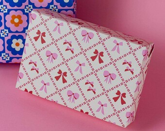 May include: A gift box wrapped in white paper with a repeating pattern of pink and red bows. The bows are set in a grid pattern with small red dots. Another box with a blue floral pattern is in the background.