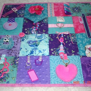 Puede incluir: Una manta sensorial multicolor con varias texturas y objetos. La manta está hecha de tela morada, rosa y azul y presenta una variedad de artículos, incluyendo una flor, un pájaro, un corazón, una llave, una cremallera y un botón.