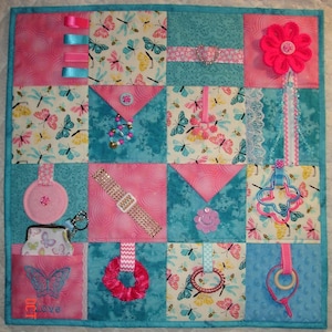 May include: A multi-coloured baby activity quilt with various textures and objects. The quilt is made of pink, blue and yellow fabric and features a variety of textures, including ribbons, buttons, zips and a small pocket. The quilt also features a variety of objects, including a flower, a butterfly, a ring and a small mirror. The quilt is perfect for babies to explore and learn about different textures and objects.