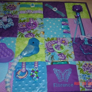 Peut inclure: Une couverture d'activité sensorielle avec un design en patchwork dans des tons de violet, bleu et vert. Elle comprend divers éléments tactiles comme un oiseau bleu, un pop-it violet en forme de cœur, des rubans, des perles et une broderie papillon avec le nom Florence et la date AUG 28 2020.