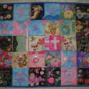 May include: A colourful quilt featuring a variety of floral and nature-themed fabric squares. Each square showcases a different design, including butterflies, hummingbirds, flowers, and inspirational quotes. The quilt is finished with a blue border and a date in the bottom right corner.
