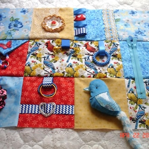May include: A handmade sensory activity mat with various textures and interactive elements. Features include a blue bird plush, a zip, ribbons, a heart-shaped embellishment, and patterned fabrics with floral and bird designs. The mat is designed for tactile exploration.