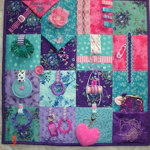 May include: A sensory quilt in shades of purple, teal, and pink, featuring a variety of textures and interactive elements. Includes a buckle, zip, ribbons, buttons, a scrunchie, a flower, a purse, and a heart-shaped keyring.