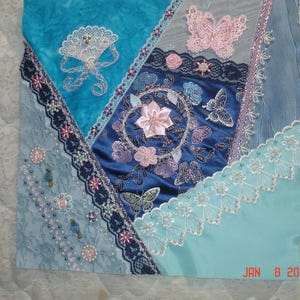 May include: A blue and white quilt block featuring a variety of embroidered designs, including butterflies, flowers, and lace. The block is made up of several different fabrics and is sewn together with a variety of stitches.