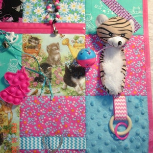 May include: A sensory board with a patchwork design, featuring a plush tiger, a zipper pocket, and a pop-it toy. The board incorporates various textures and interactive elements, with a pink border. The fabric includes floral and kitten patterns.
