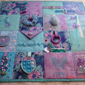 Puede incluir: Una manta de actividades sensoriales rectangular con un mosaico de telas azules, moradas y verde azulado. La manta incluye varios elementos táctiles como una goma para el pelo azul, un juguete antiestrés en forma de corazón, una cremallera, cintas, cuentas y un bolsillo bordado con el nombre Sally y un diseño de mariposa.