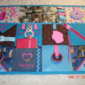May include: A blue and pink sensory activity mat with various textures and objects for babies and toddlers. The mat features a variety of textures, including soft fabrics, crinkly materials, and smooth surfaces. It also includes a variety of objects, such as a zipper, a buckle, a key chain, a teether, and a mirror. The mat is labelled "Mom" and "Mopsy".
