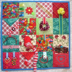 May include: A colourful baby activity quilt with various textures and elements for sensory play. It features a red bird, a flower, a zipper, a heart, a belt buckle, a beaded necklace, and the word "LOVE" embroidered on a red square.