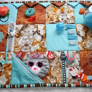 May include: A blue and brown baby activity play mat with various textures and objects for sensory stimulation. The mat features a variety of cat-themed fabrics, a zipper, a bell, a flower, a smiley face, a bow, a key chain, a stuffed toy, a teether, and a small stuffed animal.