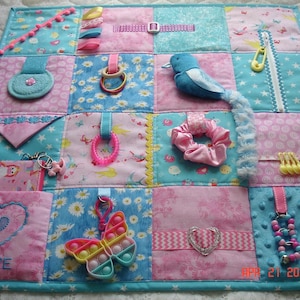 May include: A sensory activity mat with a patchwork design in blue and pink. It includes various textures and interactive elements such as a plush bird, rings, a butterfly fidget toy, and a scrunchie. The mat has the word "LOVE" embroidered on it. The mat is designed for sensory exploration.