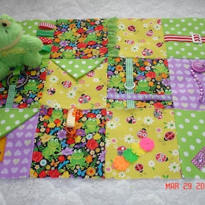 May include: A multi-coloured fabric square with various textures and patterns, including green polka dots, yellow flowers, and purple hearts. There are several small toys attached to the fabric, including a green frog, a ladybug, and a ribbon. The fabric is designed for babies and toddlers to explore and play with.