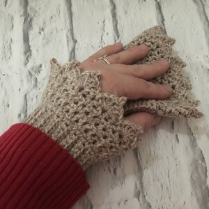 May include: Beige crocheted fingerless gloves with a scalloped edge. The gloves are worn on a hand with a ring on the finger.
