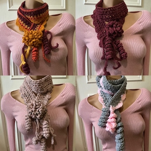 Puede incluir: Se muestran cuatro bufandas de ganchillo hechas a mano, cada una con un diseño único y elementos decorativos. Los colores incluyen naranja, burdeos, rosa y gris. Cada bufanda se muestra en un maniquí.