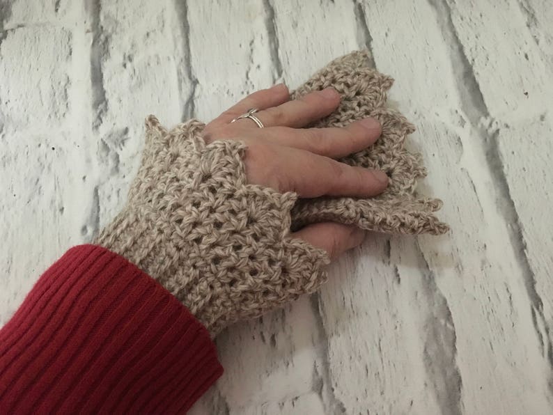 May include: Beige crocheted fingerless gloves with a scalloped edge. The gloves are worn on a hand with a ring on the finger.