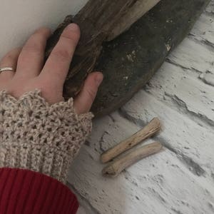 May include: A close-up of a person's hand wearing a beige crocheted wrist warmer with a scalloped edge. The hand is resting on a piece of driftwood.